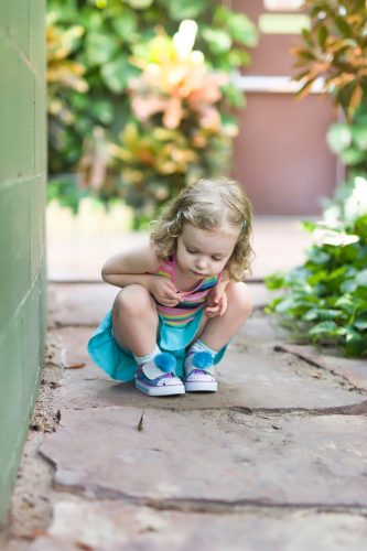 Little blonde girl squatting down with a curious expression on her face at a butterfly on the ground