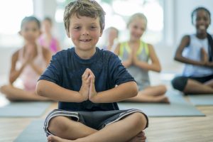 A multi-ethnic group of elementary school students are in indoors in a fitness center. They are wearing athletic clothing. They are sitting on the floor and holding a meditation pose.  A Caucasian boy is in front.